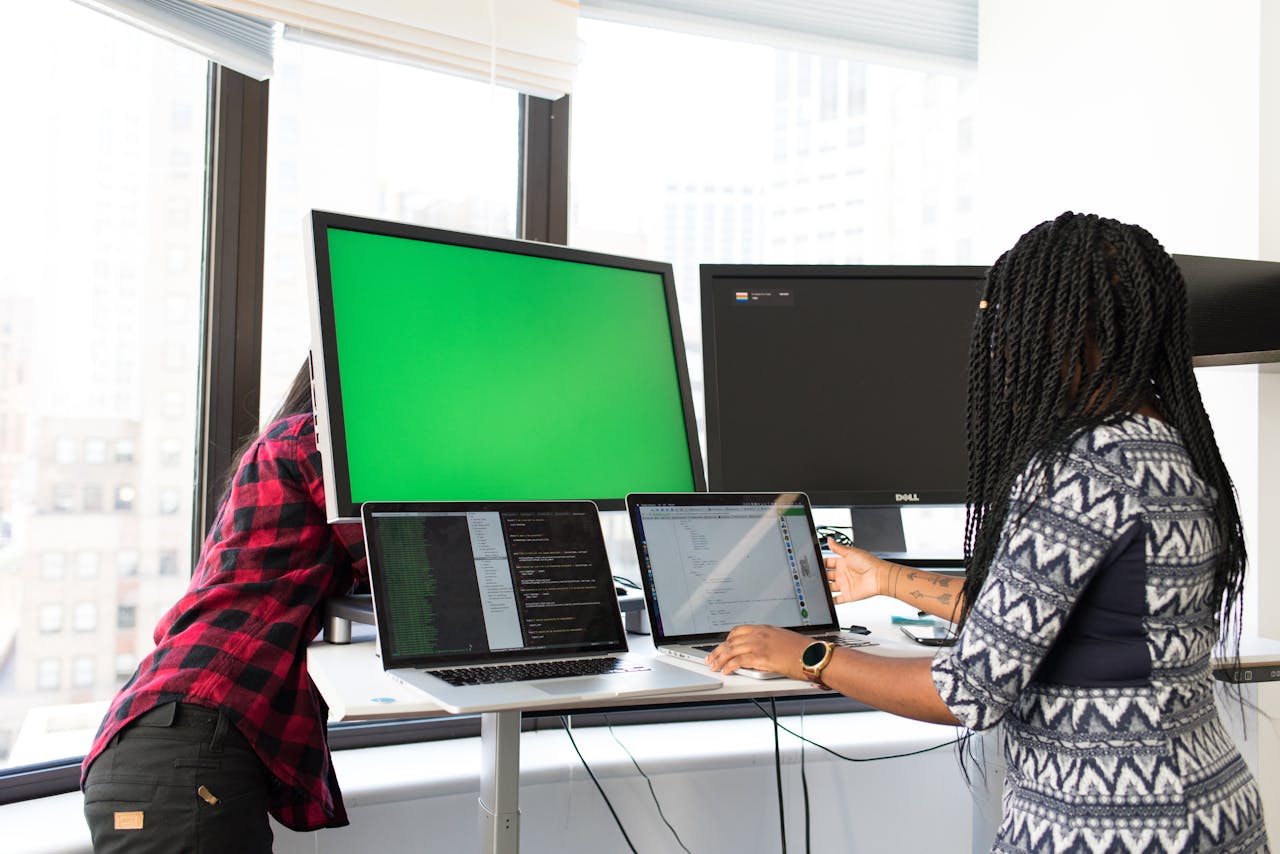 About Us Two women working on laptops and monitors in a bright office setting, focused on technology and teamwork.